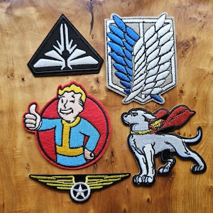 May include: A selection of embroidered patches with diverse designs. Includes a black triangle with a white design, a shield with blue and white wings, a cartoon character giving a thumbs up, a dog with a cape, and a winged emblem.
