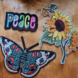 May include: Embroidered patches including a butterfly, a sunflower, and the word "Peace". The butterfly has teal, red, and blue details. The sunflower has yellow and orange petals. The word "Peace" is in colourful letters.