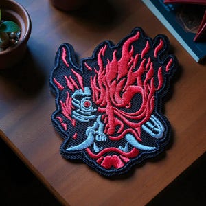 Op de afbeelding: Geborduurde patch met een gestileerd rood demonengezicht met vlammen, tegen een zwarte achtergrond. Het ontwerp bevat blauwe en witte accenten, wat een opvallend contrast creëert. Geschikt voor jassen, tassen of andere stoffen items.