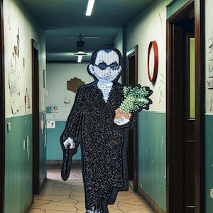 Peut inclure: Une œuvre d'art en tissu représentant une figure en long manteau noir, tenant un bouquet de plantes vertes et un objet noir, dans un couloir. La figure a un visage blanc et porte des lunettes de soleil. Le couloir a des murs verts et un sol carrelé fissuré.