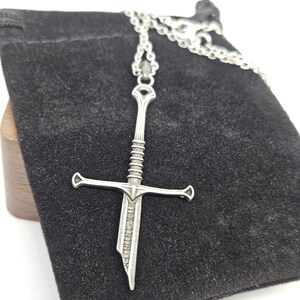 Pode incluir: Um colar com pingente de espada prateada. O pingente apresenta um design detalhado de espada com guarda-m&atilde;o e cabo texturizado. O colar &eacute; exibido em uma bolsa de veludo preta.