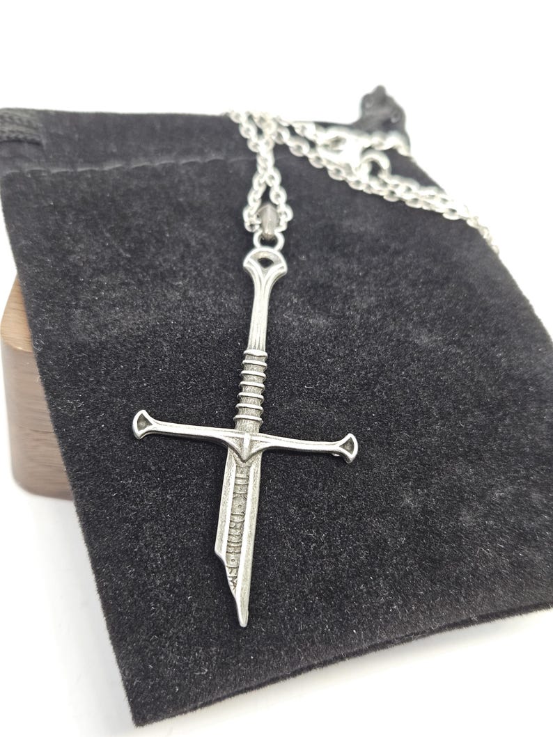 Pode incluir: Um colar com pingente de espada prateada. O pingente apresenta um design detalhado de espada com guarda-m&atilde;o e cabo texturizado. O colar &eacute; exibido em uma bolsa de veludo preta.