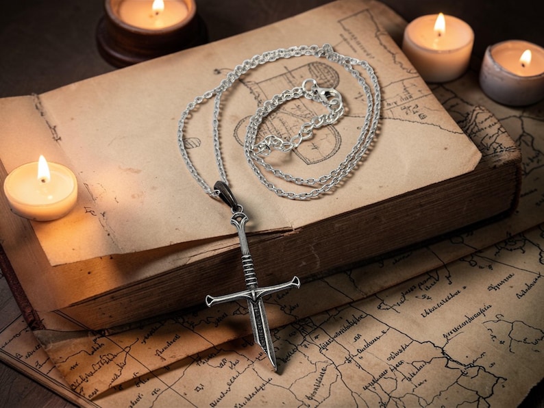Pode incluir: Um colar com pingente de espada prateada com l&acirc;mina e punho detalhados, repousando sobre um livro antigo aberto com um mapa. Velas acesas proporcionam um brilho quente, real&ccedil;ando o tema medieval ou de fantasia.