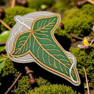 May include: Embroidered patch depicting a green leaf with gold-coloured veins and trim, set against a light grey background. The leaf design is detailed and prominent, with a white outline. The patch is likely for clothing or accessories.