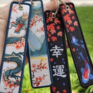 Peut inclure: Porte-clés brodés avec des motifs d'inspiration japonaise. L'un représente un dragon turquoise avec des accents rouges et des fleurs de cerisier. Un autre représente un paysage de montagne avec des fleurs de cerisier. Le troisième porte les caractères japonais pour "chance".