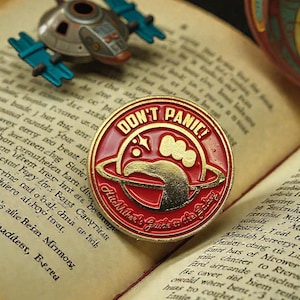 Könnte beinhalten: Rote und goldene Emaille-Anstecknadel mit der Aufschrift "DON'T PANIC!" und einem Planeten-Design. Die Anstecknadel liegt auf einem offenen Buch mit gealterten Seiten, neben einem kleinen Spielzeugraumschiff.