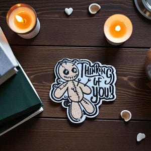 May include: An embroidered patch with a voodoo doll design and the text "Thinking of You!". The patch is beige with black stitching and pins, set against a white and light blue background. The image also includes lit candles and decorative items.
