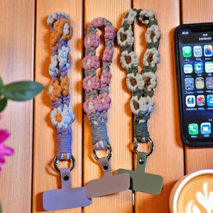 Peut inclure: Trois lanières de téléphone en macramé, violet, rose et vert, chacune avec un motif floral différent. Les lanières ont un anneau métallique et une pince de téléphone colorée. Le fond est une surface en bois.