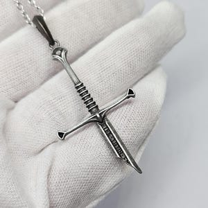 Pode incluir: Um colar com pingente prateado com um design detalhado de espada. A espada tem uma guarda em forma de cruz e um cabo texturizado. O pingente pende de uma delicada corrente prateada, criando um acess&oacute;rio marcante.