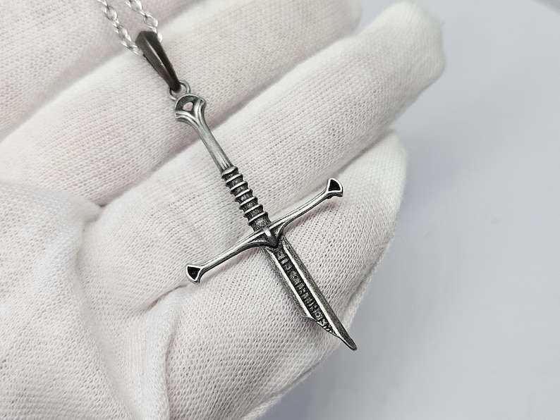 Pode incluir: Um colar com pingente prateado com um design detalhado de espada. A espada tem uma guarda em forma de cruz e um cabo texturizado. O pingente pende de uma delicada corrente prateada, criando um acess&oacute;rio marcante.