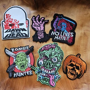 May include: A collection of embroidered patches with horror themes. Designs include a zombie hand, a pumpkin skull, and a zombie with exposed brain. Text includes "Dawn of the Dead", "No Lives Matter", and "Zombie Hunter".