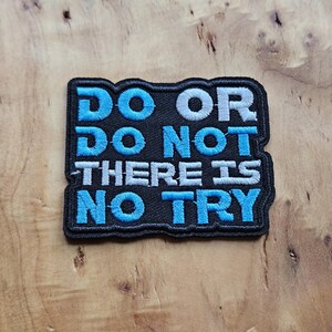 Op de afbeelding: Zwart geborduurde patch met de tekst "DO OR DO NOT THERE IS NO TRY" in blauwe en witte letters. De patch heeft een zwarte rand en is geplaatst tegen een houten achtergrond. Een motiverende quote.
