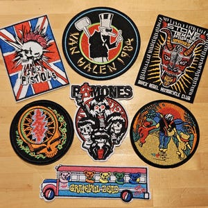 Könnte beinhalten: Eine Sammlung von aufgestickten Aufnähern mit Bandlogos und Punkrock-Motiven. Designs umfassen einen Totenkopf mit Union Jack, einen Teufel und einen Bus mit Bären. Die Aufnäher sind in verschiedenen Formen und Größen mit leuchtenden Farben.