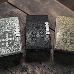 May include: Three rectangular metal lighters in silver, matte black, and antique brass finishes. Each lighter features an intricate embossed Saint Benedict medal design on the front, with a vertical ribbed ignition roller on the right side.