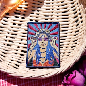 May include: Embroidered patch depicting a woman with sunglasses, a floral crown, and blue hair. The patch has a blue background with red sun rays. The patch is rectangular with a black border. The patch is on a wicker basket.