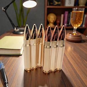 May include: Two sets of gold-coloured bullet-shaped lighters with copper-coloured tips are displayed on a wooden surface. A pen and a book are also visible. The background includes a lamp, a glass of amber liquid, and a bookshelf.