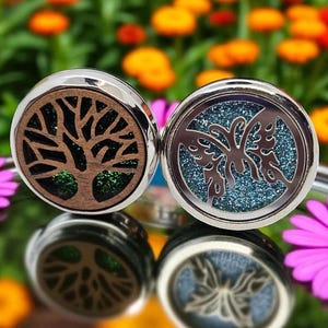 May include: Two round aromatherapy diffuser pendants with silver rims. One has a tree design with brown wood grain and green accents. The other features a butterfly design with a blue glitter background. The pendants are on a reflective surface.