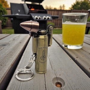 Puede incluir: Un encendedor con forma de bala de color latón con una punta de color cobre, un llavero y el texto "HUNTER BULLET LIGHTER". Un vaso de líquido amarillo está junto al encendedor sobre una mesa de madera. Una parrilla está en el fondo.