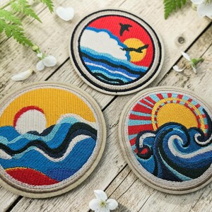 以下が含まれることがあります： 海と太陽のデザインが施された3つの円形の刺繍ワッペン。1つは赤い空に黄色い太陽と鳥、もう1つは青い波の上に黄色い太陽、3つ目は大きな波の上に赤い光線を放つ黄色い太陽が描かれています。これらはすべて円形の刺繍パッチです。