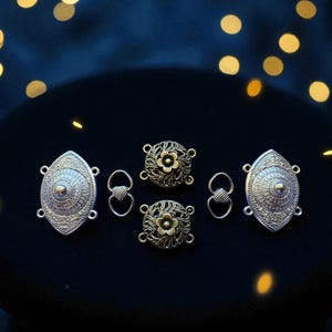 May include: A collection of decorative metal jewellery findings. The image shows oval clasps with intricate patterns and floral connectors in silver and bronze hues. The items are arranged on a dark surface with bokeh lights.