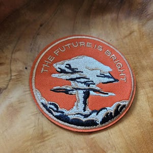 May include: Circular orange embroidered patch with the words "THE FUTURE IS BRIGHT". The patch features a stylized mushroom cloud design in white, black, and grey.