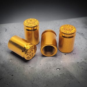 Puede incluir: Cuatro objetos metálicos dorados, que se asemejan a casquillos de bala, están dispuestos sobre una superficie gris texturizada. Los objetos tienen forma cilíndrica con una parte superior detallada y un interior roscado. La iluminación crea reflejos y sombras.
