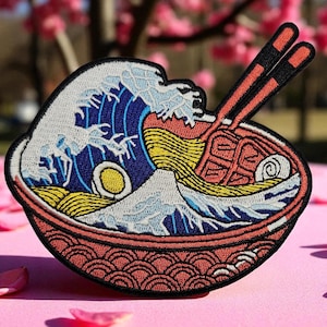 May include: Embroidered patch depicting a bowl of ramen with chopsticks. The design incorporates a wave, mountain, and egg, in red, blue, yellow, and white. A decorative item.