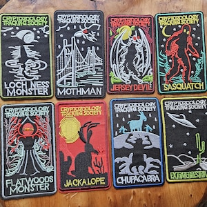 Pode incluir: Uma coleção de oito patches bordados com ilustrações de criptídeos. Cada patch tem uma borda preta e o texto "Cryptozoology Tracking Society". Os desenhos incluem o Monstro do Lago Ness, Mothman, Jersey Devil e Sasquatch. Os patches estão em uma superfície de madeira.