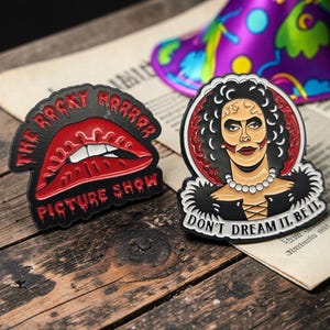 May include: Two enamel pins featuring The Rocky Horror Picture Show. One pin displays red lips with the text THE ROCKY HORROR PICTURE SHOW. The other pin depicts a character with dark hair, a pearl necklace, and the text DON'T DREAM IT. BE IT.