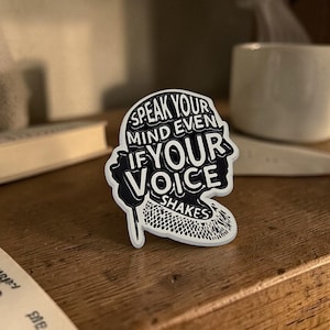Peut inclure: Un autocollant noir et blanc en forme de silhouette d'une personne, avec le texte "SPEAK YOUR MIND EVEN IF YOUR VOICE SHAKES". L'autocollant est posé sur une surface en bois.