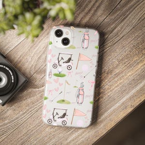 May include: A clear phone case with a golf-themed pattern. The design includes pink golf carts, flags, golf bags, golf balls, and hearts on a light gray background. The case is on a wooden surface.