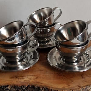 May include: Set of six stainless steel coffee cups and saucers. The cups have a classic design with a rounded bowl and a curved handle. The saucers are round with a slightly raised edge. The set is displayed on a wooden surface.