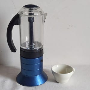 May include: A blue and clear coffee maker with a black handle and a white coffee cup. The coffee maker has a transparent top and a blue base. The cup is small and white, placed next to the coffee maker.