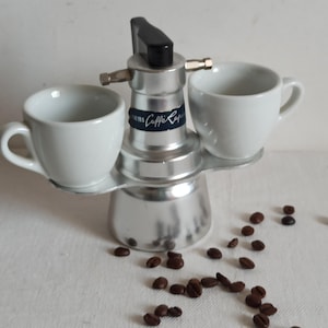 Puede incluir: Una cafetera espresso plateada con dos tazas de cerámica blancas. La cafetera espresso tiene un asa negra y el texto "Caffe Rapido". Alrededor de la base hay varios granos de café.