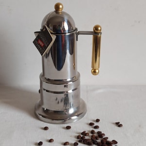 May include: A stainless steel stovetop espresso maker with a gold-colored handle and top. A small tag is attached to the side. Coffee beans are scattered in front of the espresso maker.