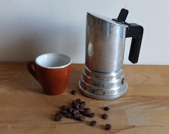 May include: A silver and black stovetop espresso maker with a brown ceramic cup and coffee beans scattered on a wooden surface.