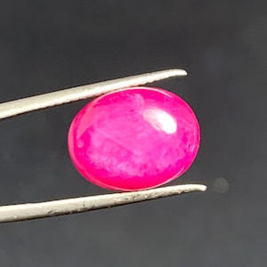 Könnte beinhalten: Ein leuchtend pinkfarbener, runder Edelstein wird von einer silbernen Pinzette vor einem dunklen Hintergrund gehalten. Der polierte Stein hat eine glatte Oberfläche mit inneren Lichtreflexen, die seine Farbe und Form zeigen.