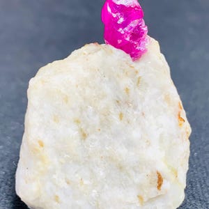 May include: A vivid, hot pink ruby crystal sits on a rough, off-white stone. The ruby's crystalline structure is visible, reflecting light. The stone has a textured surface with small, brown flecks. This image showcases a natural gemstone.