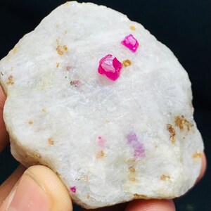 Pode incluir: Uma pedra branca áspera com vários pequenos cristais de rubi rosa brilhante embutidos. A pedra tem uma textura ligeiramente áspera e algumas áreas com manchas castanhas e douradas.