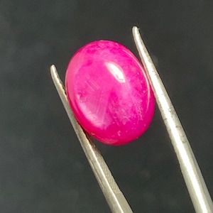 May include: A vivid, oval-shaped, hot pink gemstone is held by silver tweezers against a dark backdrop. The polished stone has a smooth surface and a slight sheen, showcasing its rich colour.