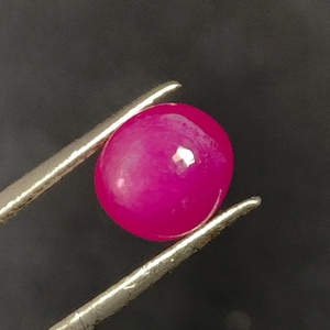 May include: A vibrant, polished, round, hot pink gemstone is held by silver tweezers against a dark background. The gemstone has a smooth, reflective surface, and is a deep, saturated shade of pink.