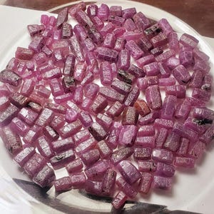 以下が含まれることがあります： 未加工のルビーの原石のコレクション。原石は濃いピンクがかった赤色で、表面はざらざらとした質感です。白い表面に散らばっており、自然の美しさを視覚的に表現しています。ルビーはサイズと形状が様々です。