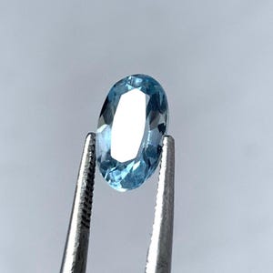 May include: An oval-shaped, light blue gemstone held by silver tweezers. The gemstone's facets reflect light, creating a sparkling effect. The background is a soft, neutral grey.