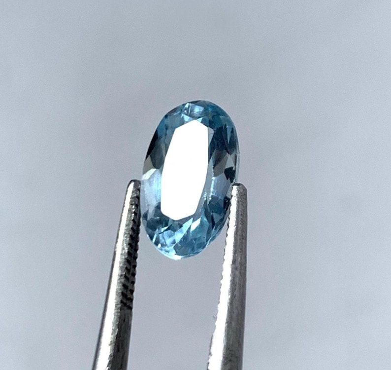 May include: An oval-shaped, light blue gemstone held by silver tweezers. The gemstone's facets reflect light, creating a sparkling effect. The background is a soft, neutral grey.