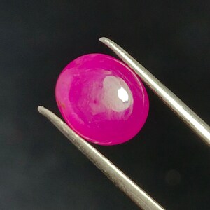 Peut inclure: Une pierre précieuse rose vif, de forme ovale, est maintenue par une pince argentée sur un fond noir. La pierre polie reflète la lumière, révélant sa surface lisse et sa couleur riche. Il s'agit probablement d'un rubis.