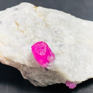 May include: A rough, white rock formation with a vibrant, hot pink crystal protruding from its surface. A smaller pink crystal is visible at the bottom right. The rock has a textured appearance, and the crystals are sharply defined.