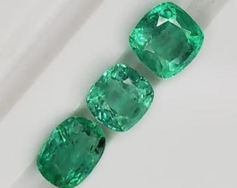 May include: Three vibrant green emerald gemstones with a cushion cut. The gems are displayed on a white surface, showcasing their clarity and brilliance. The emeralds are arranged diagonally, highlighting their individual facets.