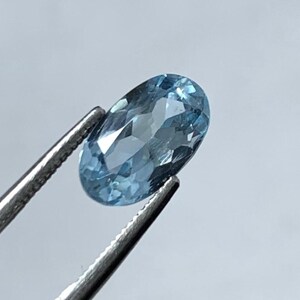 May include: A light blue, oval gemstone is held by silver tweezers. The gem's surface is faceted, catching the light and creating a sparkling effect. The background is a soft, neutral colour, emphasising the gem's clarity and colour.