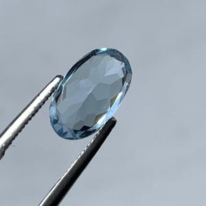 May include: A light blue, oval-cut gemstone held by silver tweezers. The gemstone's facets catch the light, creating a sparkling effect. The background is a plain, pale grey.