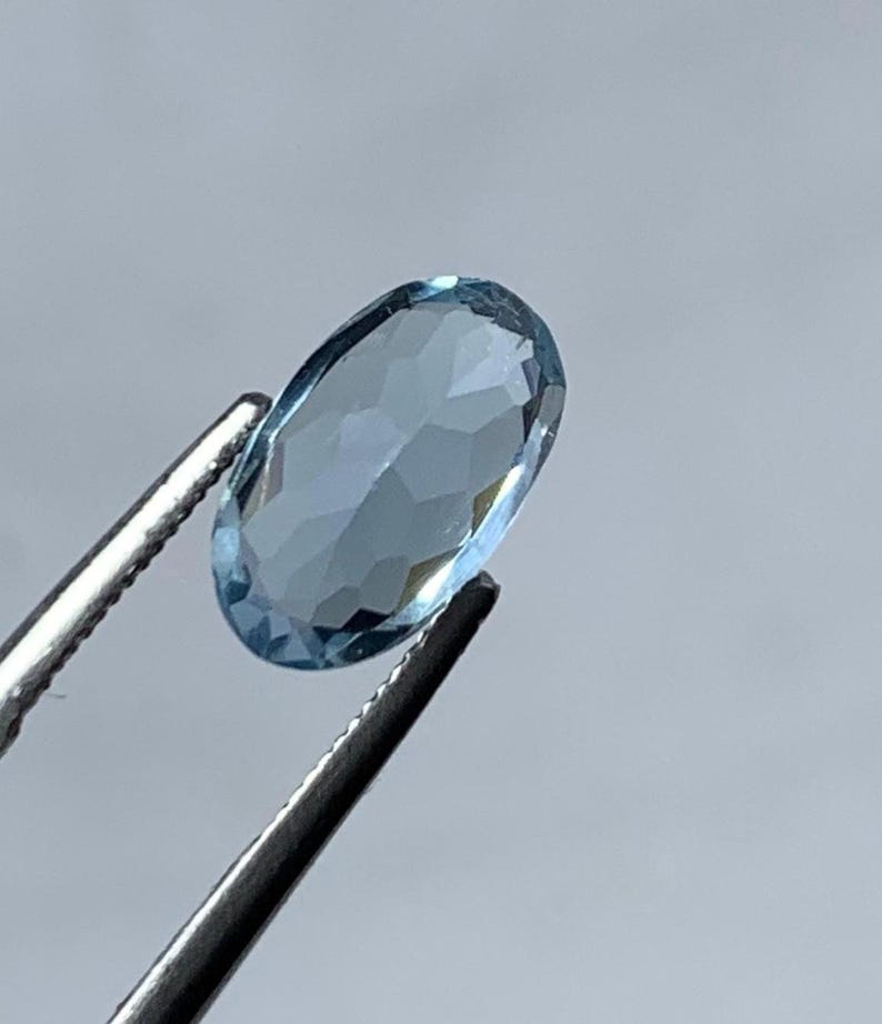May include: A light blue, oval-cut gemstone held by silver tweezers. The gemstone's facets catch the light, creating a sparkling effect. The background is a plain, pale grey.